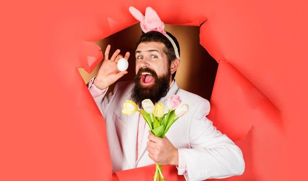 Пасхальный день. Счастливый бородатый мужчина с пасхальным яйцом и цветами. Кролик смотрит в бумажное отверстие. - Фото, изображение