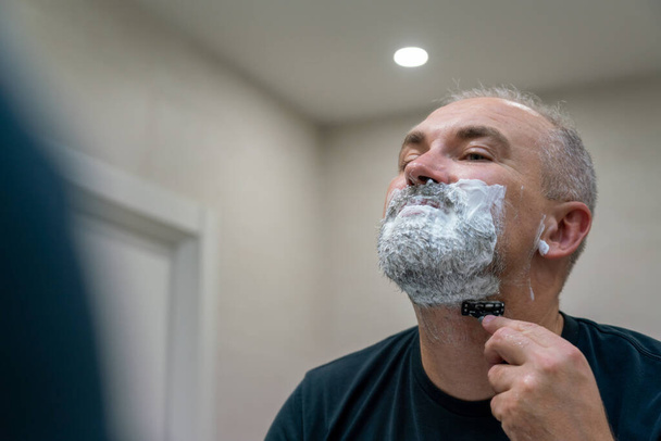 Maduro hombre de pelo gris restyling su barba a sí mismo en casa usando navaja de afeitar - Foto, imagen