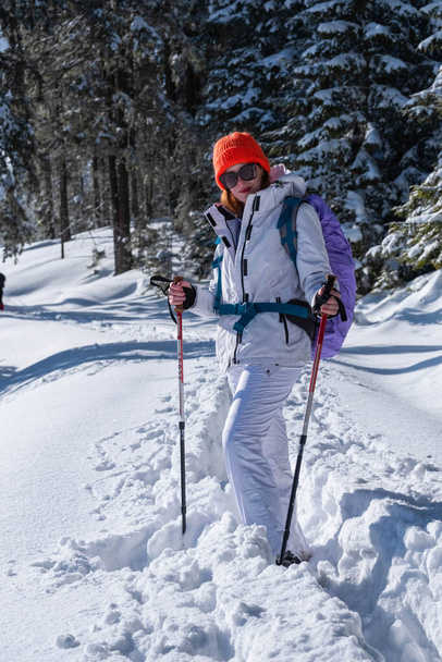 Portrait of a young white caucasian female hiker dressed in white posing for the camera in sunglasses. Winter season and the sun shining in the background - Photo, Image