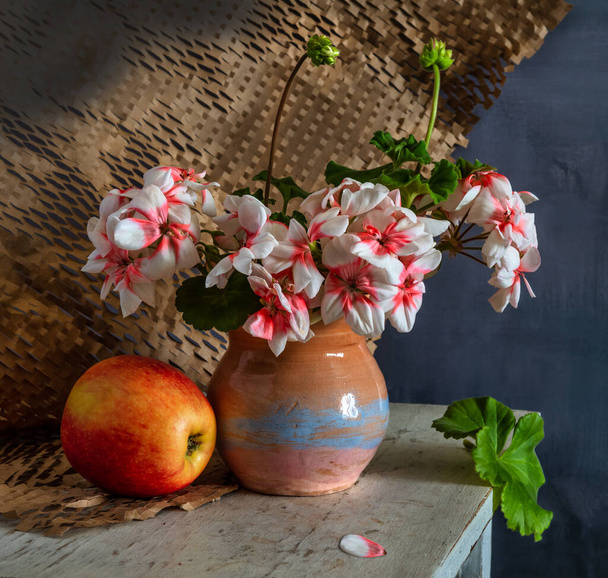 Bouquet of blooming geraniums in a ceramic vase on a dark background. and a ripe apple. - Photo, Image