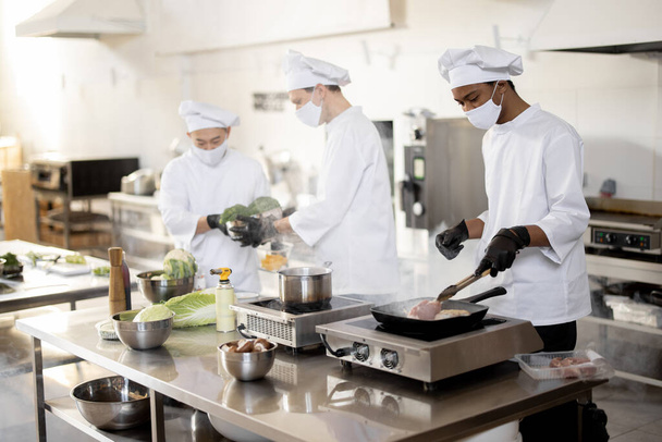 Wielorasowy zespół kucharzy w mundurach i maskach do twarzy gotowanie posiłków dla restauracji w kuchni - Zdjęcie, obraz