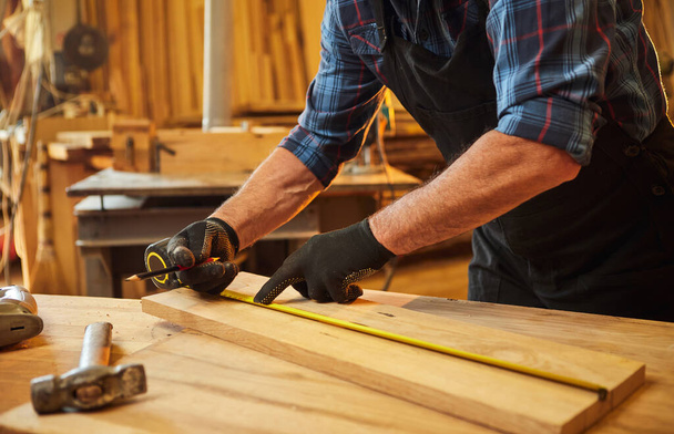 Carpintero trabajando con una madera, marcando tablón con un lápiz y tomando medidas para cortar un trozo de madera para hacer un mueble en un taller de carpintería, vista de cerca - Foto, Imagen