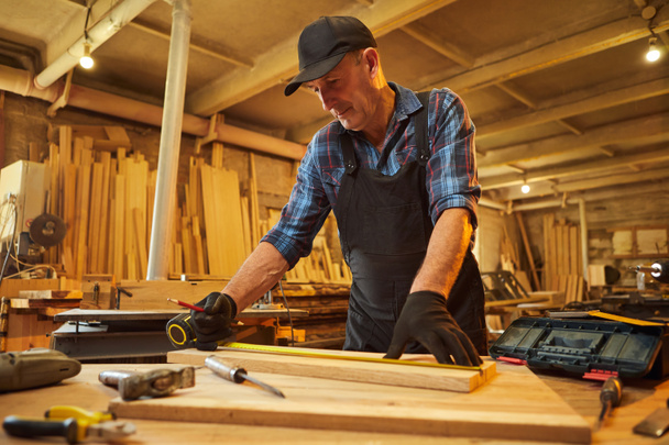 Retrato de un carpintero senior trabajando con una madera, marcando tablón con un lápiz y tomando medidas para cortar un trozo de madera para hacer un mueble en el taller de carpintería - Foto, Imagen
