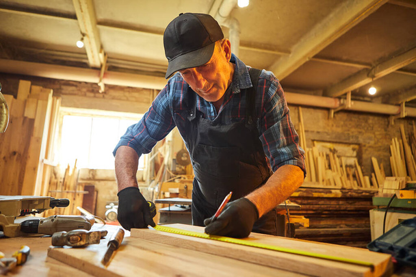 Retrato de un carpintero senior trabajando con una madera, marcando tablón con un lápiz y tomando medidas para cortar un trozo de madera para hacer un mueble en el taller de carpintería - Foto, imagen