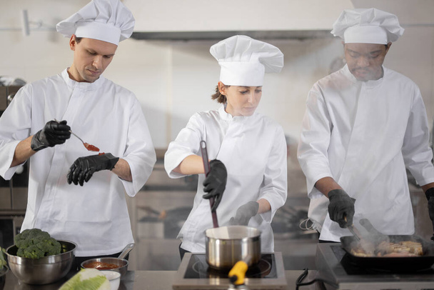 Πολυφυλετική ομάδα τριών μαγείρων που μαγειρεύουν στην επαγγελματική κουζίνα - Φωτογραφία, εικόνα