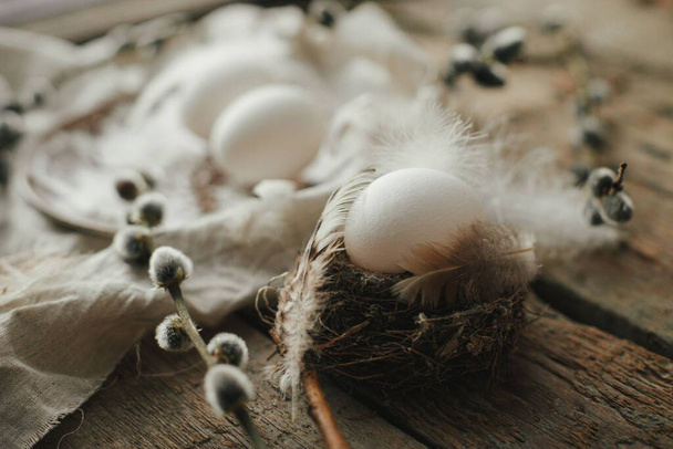 Rustic Easter still life. Natural egg in nest with feathers, vintage plate,  pussy willow branches and napkin on aged wood. Easter table decoration. Moody image - Valokuva, kuva