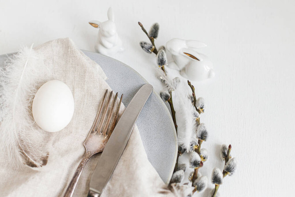 Stylish Easter table setting. Natural easter egg, pussy willow branches and feathers on modern plate, napkin and cutlery on white wooden table. Modern Easter table decoration - Foto, immagini