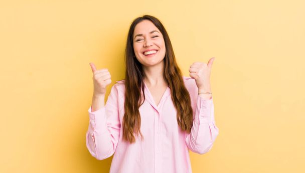 pretty caucasian woman smiling joyfully and looking happy, feeling carefree and positive with both thumbs up - Foto, Bild