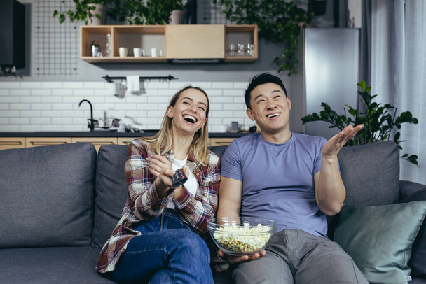Young couple in love man and woman watching TV together at home, rejoicing and eating popcorn, happy multiracial family in a new apartment - Fotografie, Obrázek