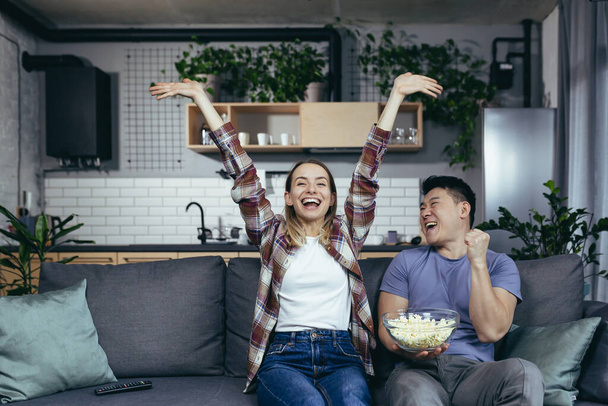 Young couple in love man and woman watching TV together at home, rejoicing and eating popcorn, happy multiracial family in a new apartment - Фото, изображение