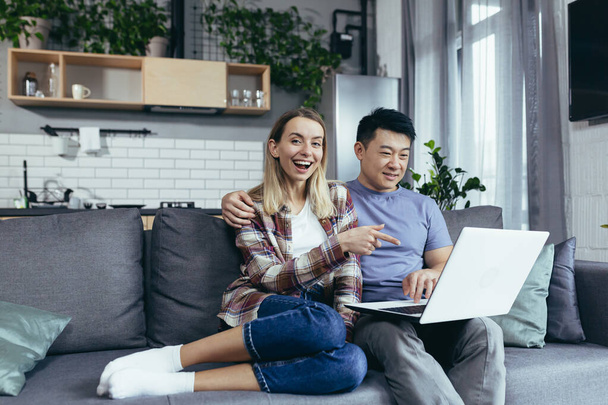 Young couple Asian man and woman at home sitting on sofa together using laptop, happy and smiling multiracial family looking at camera - Фото, зображення