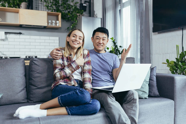 Young couple Asian man and woman at home sitting on sofa together using laptop, happy and smiling multiracial family looking at camera - Fotografie, Obrázek