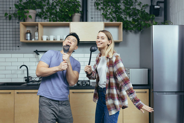 Happy young Asian man and woman couple singing and dancing together in the kitchen, having fun together, family in love - Fotografie, Obrázek