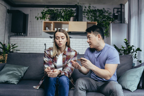 Asian man quarrels with his wife, accuses him of squandering family money, family quarrel, couple sitting on sofa, husband points to reduction of credit card account - Fotografie, Obrázek