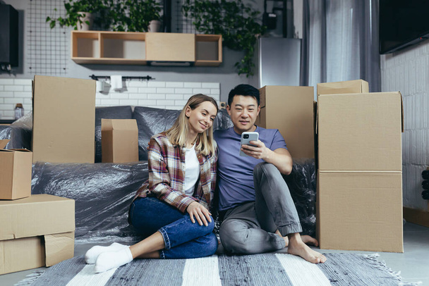 Uma jovem família em um novo apartamento entre as caixas de papelão sentado no chão e felizmente escolher nova loja on-line de móveis, use o aplicativo de telefone para fazer compras - Foto, Imagem
