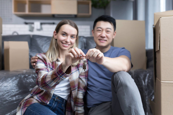 Portrait of a young couple man and woman looking at the camera and smiling holding the keys to a new apartment, multiracial family sitting on the bed of a new house hugging - Foto, Imagen