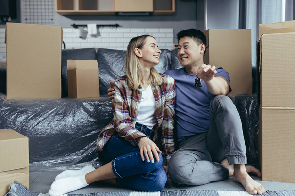 Close-up photo of a young family in a new apartment holding the keys to the house, a multiracial couple sitting in the living room among the cardboard boxes, settled in a new apartment - Foto, Bild