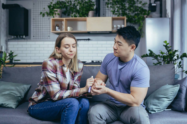 Man Asian supports wife, disappointing pregnancy test result, young multiracial couple sitting on sofa, frustrated - 写真・画像