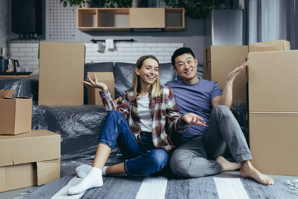 Young multiracial family man and woman sitting on the floor among cardboard boxes, looking at the camera and smiling waving hands greeting gesture - Фото, изображение