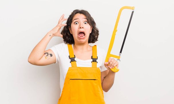 pretty hispanic girl screaming with hands up in the air. handyman and saw concept - Photo, Image