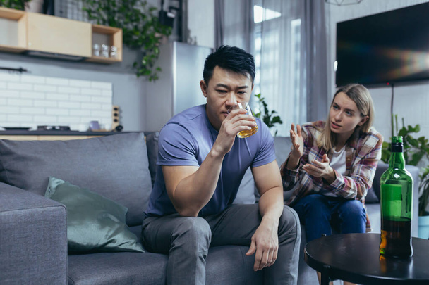 Asian husband is an alcoholic, drinks strong alcohol at home, his wife quarrels with him and tries to interfere, multiracial family at home conflict - Photo, Image