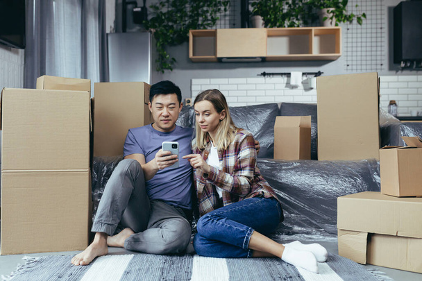 Een jong gezin in een nieuw appartement tussen de kartonnen dozen zitten op de vloer en gelukkig kiezen van nieuwe meubels online winkel, gebruik de telefoon app om te winkelen - Foto, afbeelding