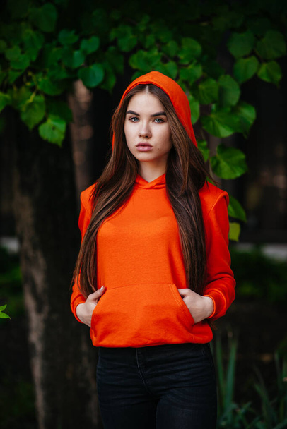 Outdoor image of a attractive young woman smiling, wearing orange knitted sweater posing on fall nature background. Beautiful female with closed eyes has dreamy expression, resting outdoor in the park - Фото, изображение