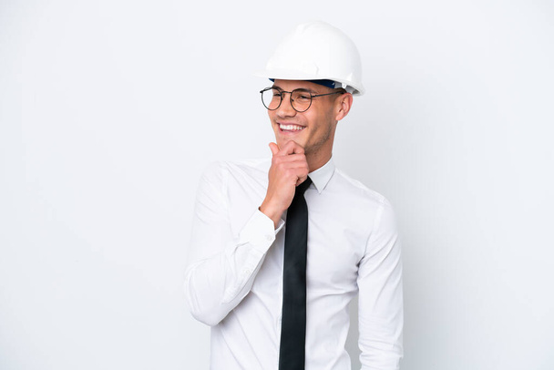 Mladý architekt kavkazský muž s helmou a drží plány izolované na bílém pozadí dívá do strany a usmívá se - Fotografie, Obrázek