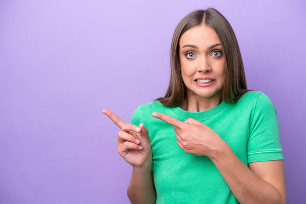 Young caucasian woman isolated on purple background frightened and pointing to the side - Photo, Image