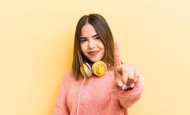 pretty hispanic girl smiling and looking friendly, showing number one. headphones concept - Foto, Imagem