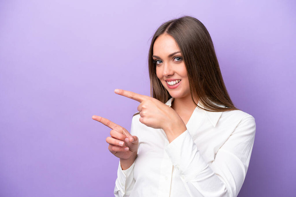 Giovane donna caucasica isolata su sfondo viola che punta il dito verso il lato e presenta un prodotto - Foto, immagini