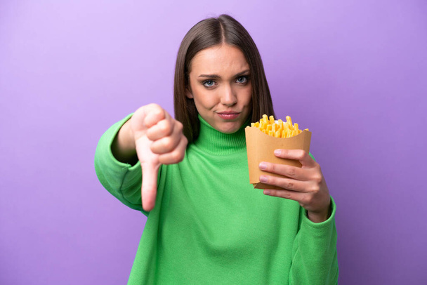 Молода біла жінка тримає смажені чіпси на фіолетовому фоні, показуючи великий палець вниз з негативним виразом
 - Фото, зображення