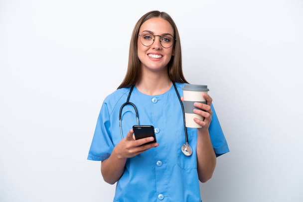 Young nurse caucasian woman isolated on white background holding coffee to take away and a mobile - Valokuva, kuva