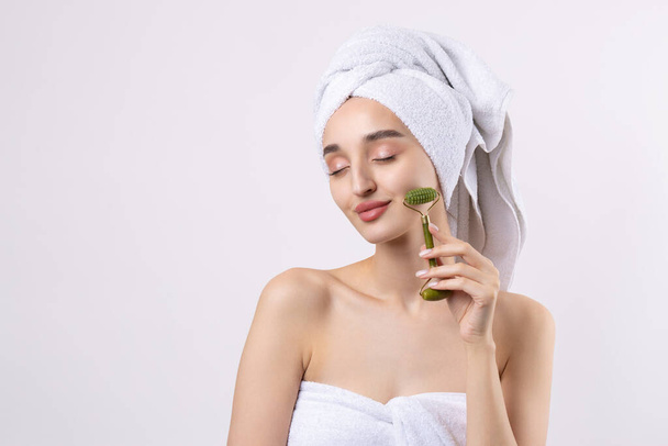 Beautiful girl with thick eyebrows and perfect skin at white background, towel on head, beauty photo. Holds a cosmetic roller. - Foto, afbeelding
