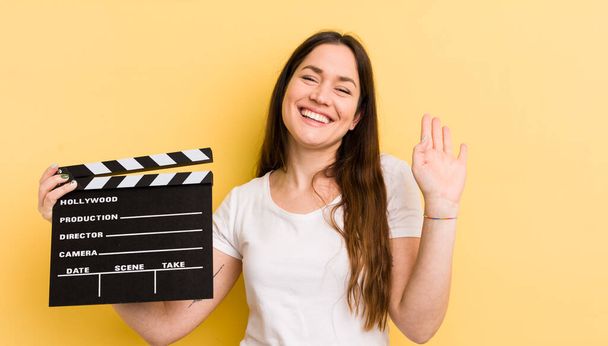 young pretty woman smiling happily, waving hand, welcoming and greeting you. cinema clapper concept - Foto, Bild
