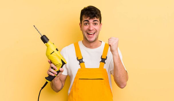 young handsome man feeling shocked,laughing and celebrating success. handyman and drill concept - Fotoğraf, Görsel