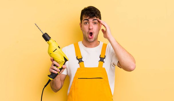 young handsome man looking happy, astonished and surprised. handyman and drill concept - Photo, Image