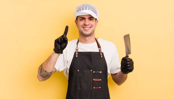 young handsome man smiling and looking friendly, showing number one. butcher concept - Фото, изображение