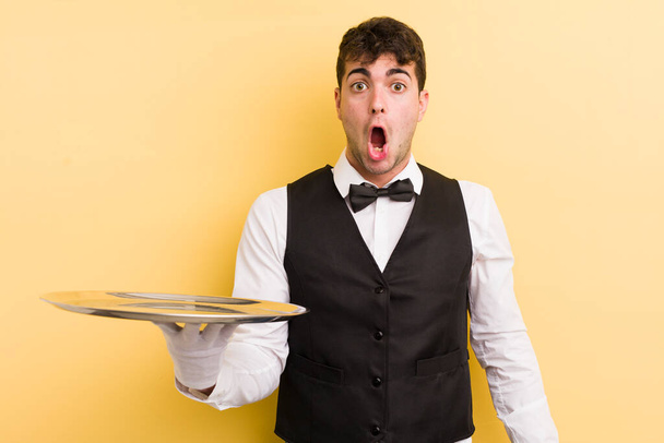 young handsome man looking very shocked or surprised. waiter and tray concept - Foto, Imagen