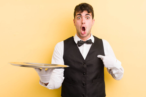young handsome man looking shocked and surprised with mouth wide open, pointing to self. waiter and tray concept - Zdjęcie, obraz