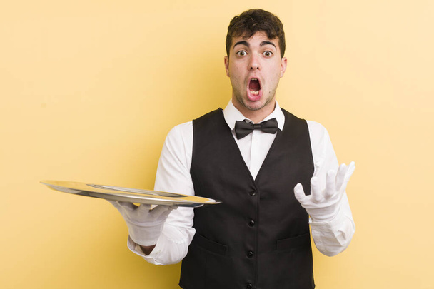young handsome man feeling extremely shocked and surprised. waiter and tray concept - Foto, imagen