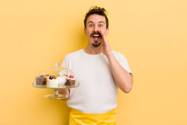 young handsome man feeling happy,giving a big shout out with hands next to mouth. home made cakes concept - Foto, imagen