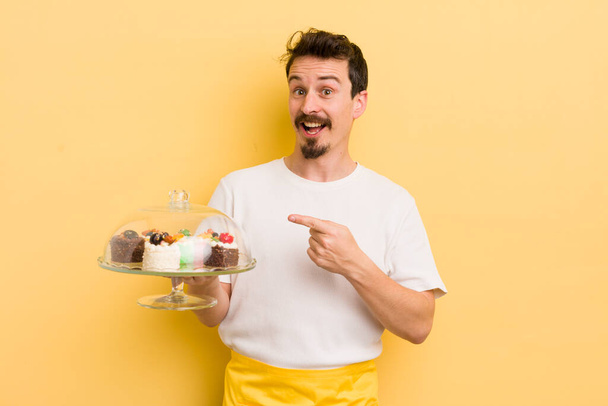 young handsome man looking excited and surprised pointing to the side. home made cakes concept - Zdjęcie, obraz
