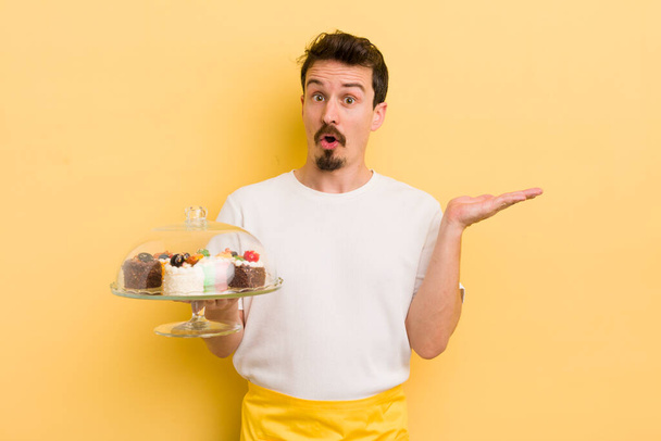 young handsome man looking surprised and shocked, with jaw dropped holding an object. home made cakes concept - Zdjęcie, obraz