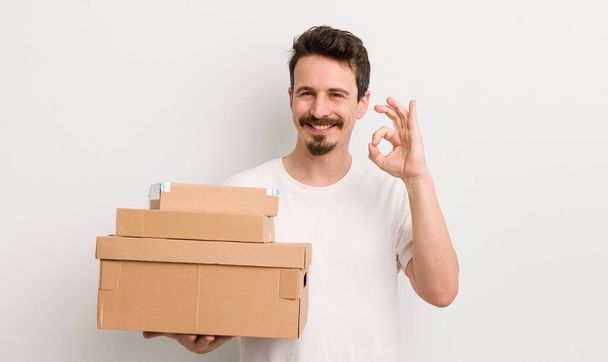 young handsome man feeling happy, showing approval with okay gesture. shipping boxes concept - Foto, Imagem