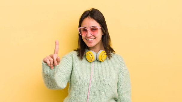 young hispanic woman smiling proudly and confidently making number one. headphones and sunglasses concept - Фото, зображення