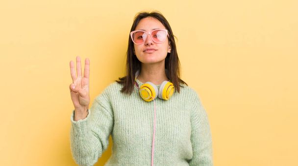 young hispanic woman smiling and looking friendly, showing number three. headphones and sunglasses concept - Фото, зображення