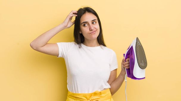 young hispanic woman feeling puzzled and confused, scratching head. clothes iron concept - Photo, Image