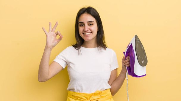 young hispanic woman feeling happy, showing approval with okay gesture. clothes iron concept - Photo, Image