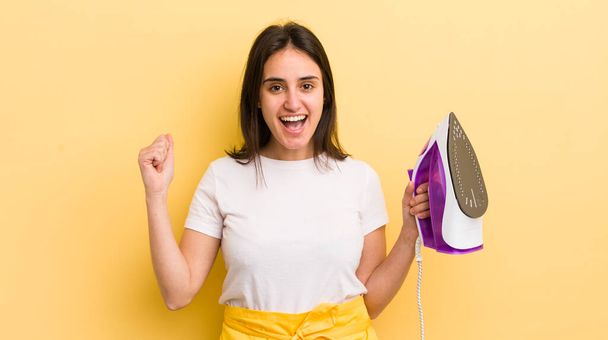 young hispanic woman feeling shocked,laughing and celebrating success. clothes iron concept - Foto, afbeelding